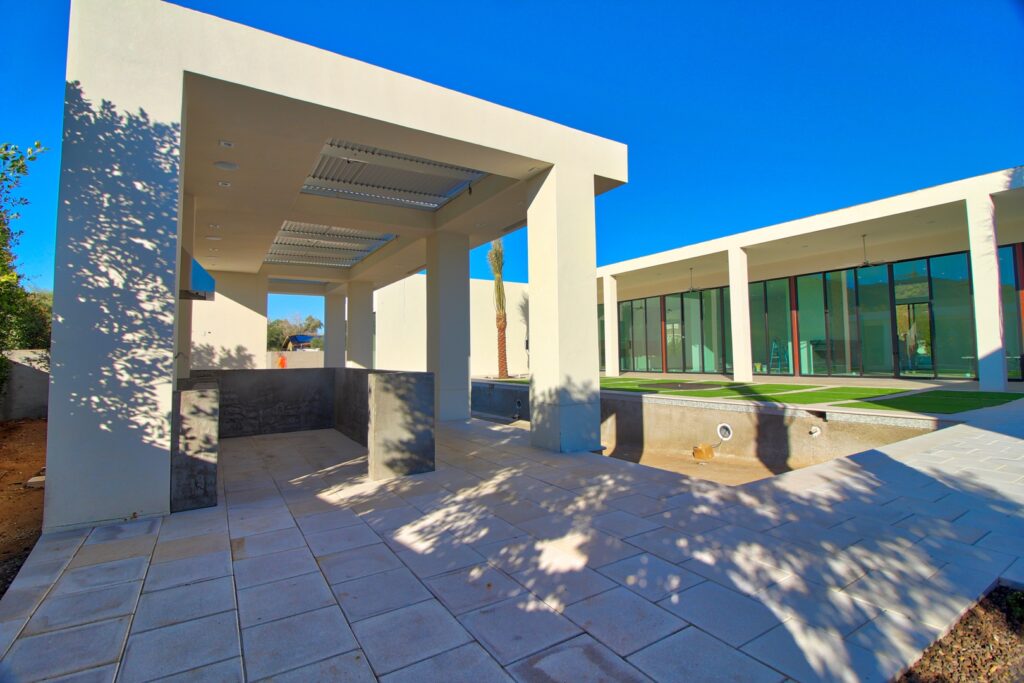 Modern outdoor patio area with pergola structure, tiled flooring, and view of a contemporary building with large windows on a clear sunny day.