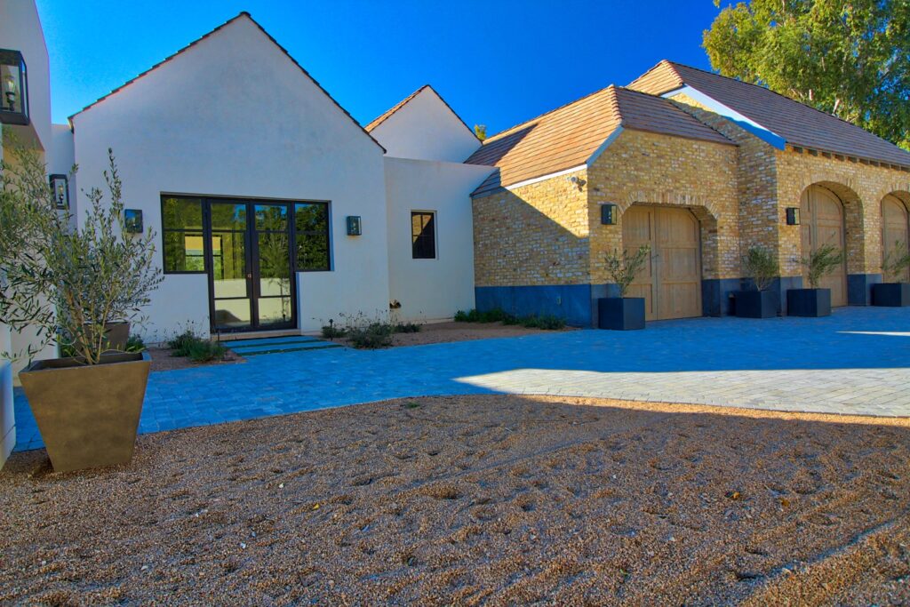 Modern house exterior with white walls, light brick accents, arched wooden garage doors, large windows, potted plants, and a gravel driveway under a clear blue sky.