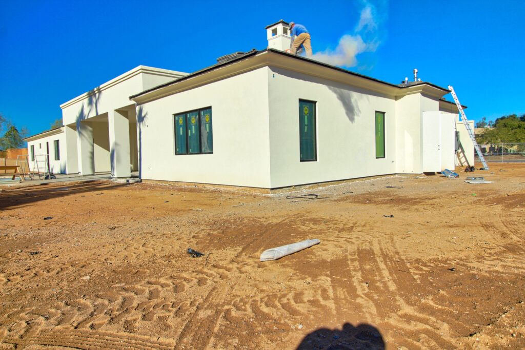 A new, single-story white house with green-trimmed windows is under construction, surrounded by bare dirt and construction materials under a clear blue sky.
