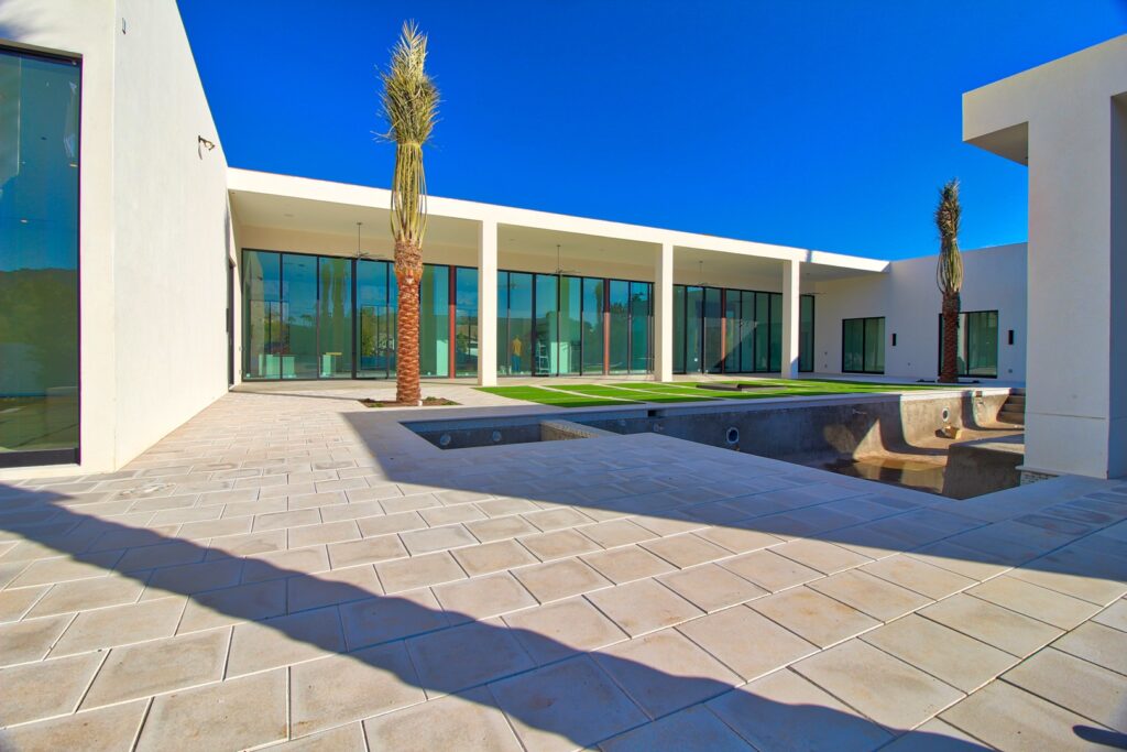 Modern courtyard of a white contemporary building with large glass doors, two tall palm trees, tiled flooring, and a small unfinished pool area under a clear blue sky.