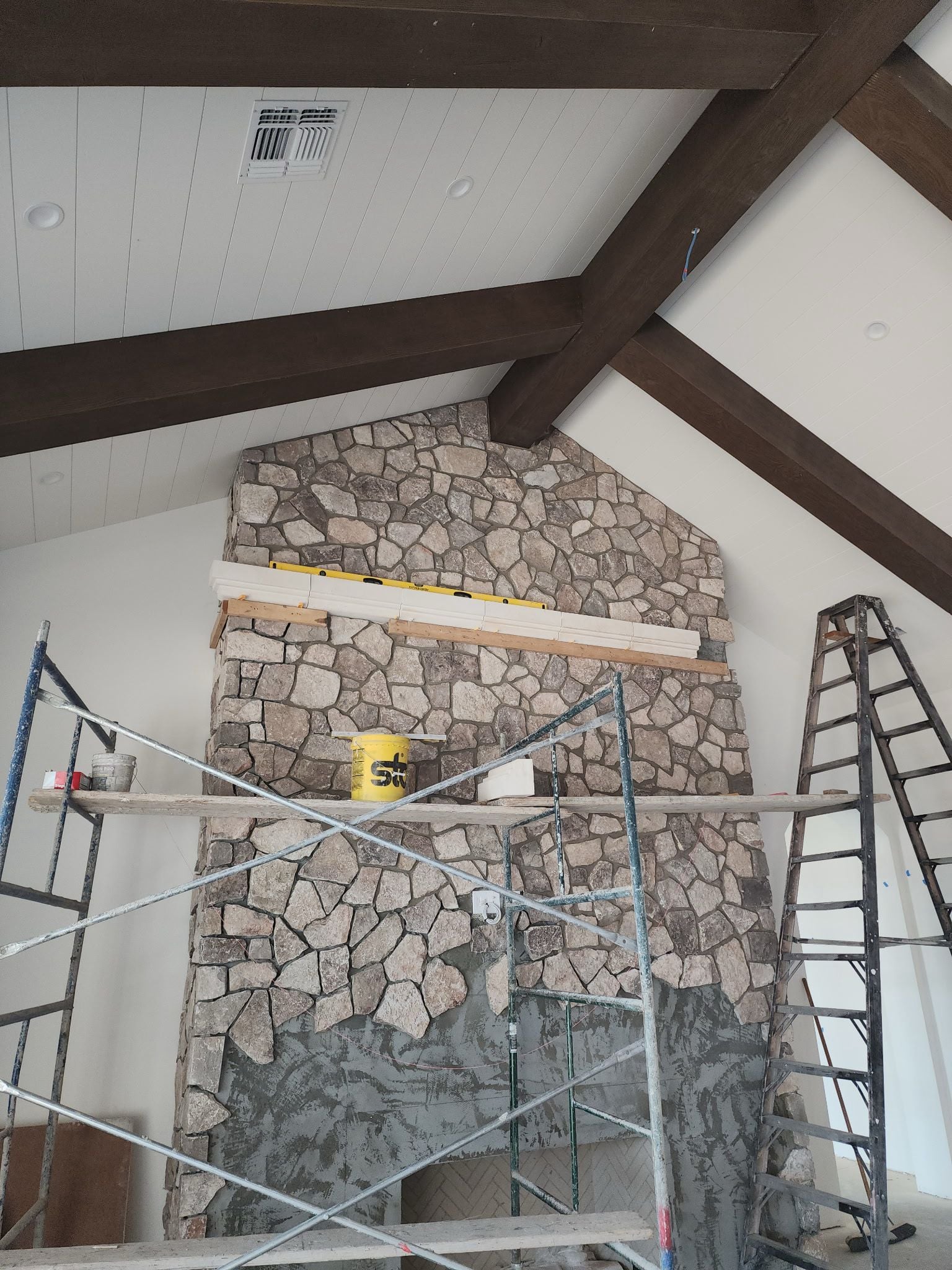 Stone fireplace under construction with scaffolding, a yellow bucket, and ladders in front; architectural elements like wood beams and a white-paneled ceiling are visible above.