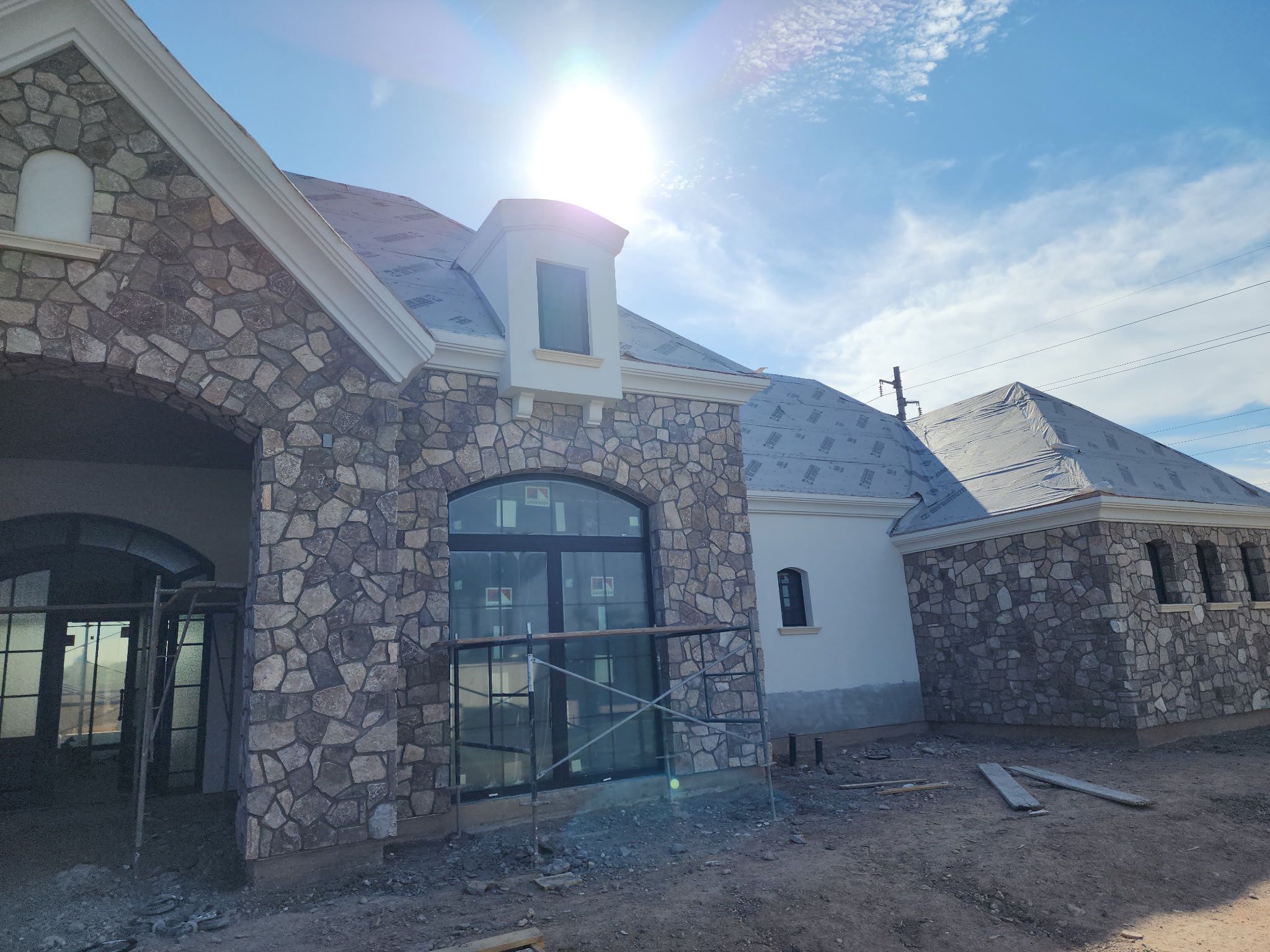 A stone house under construction featuring elegant architectural elements like arched windows and scaffolding, sunlight shining over the roof, and construction materials scattered on the ground.
